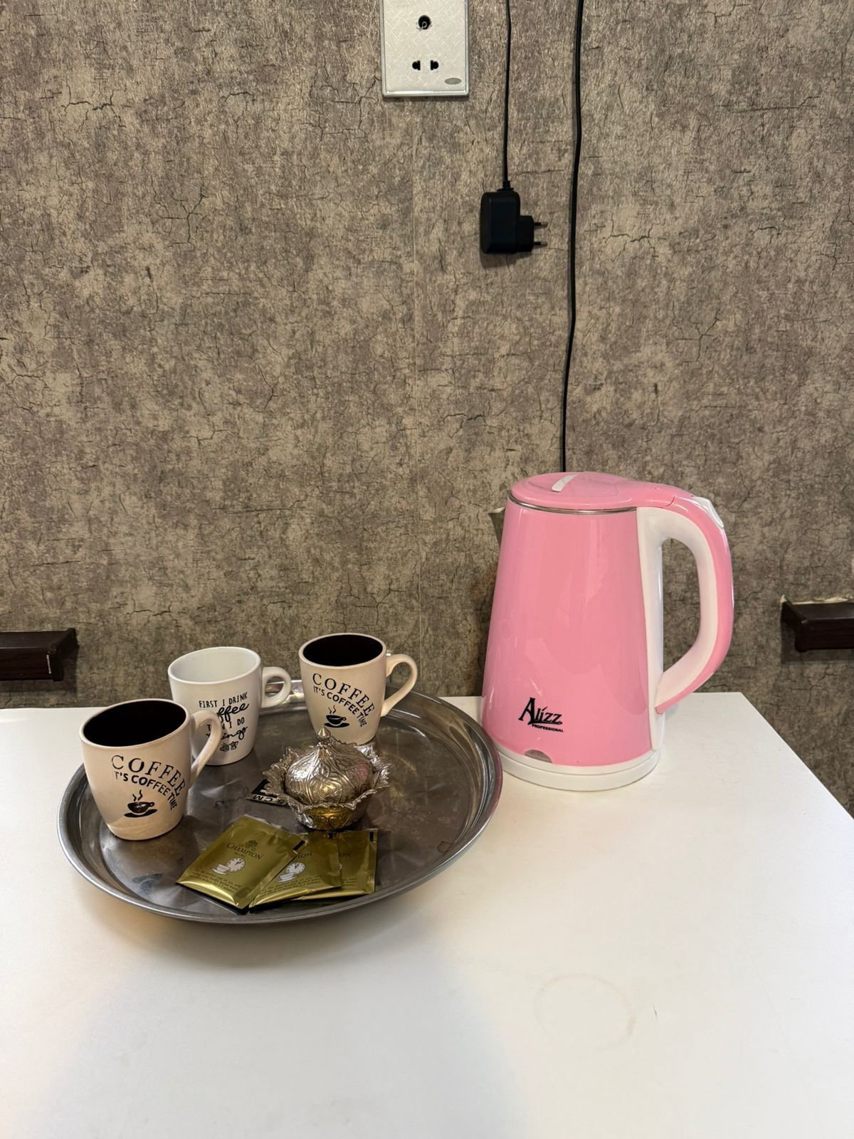 Ein Tablett mit Kaffeetassen, einer pinken Kaffeemaschine und einer Teeverpackung auf einem Tisch vor einer strukturierten Wand mit einer Steckdose.