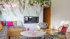 Un salon avec un tapis à motifs zébrés, une table basse ronde sur laquelle repose un livre rose, un canapé agrémenté de coussins colorés et un téléviseur fixé au mur.
