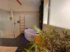 Une salle de bain moderne avec une douche à l’italienne en verre, une baignoire blanche dotée de lumières LED, une échelle en bois et une plante en pot.