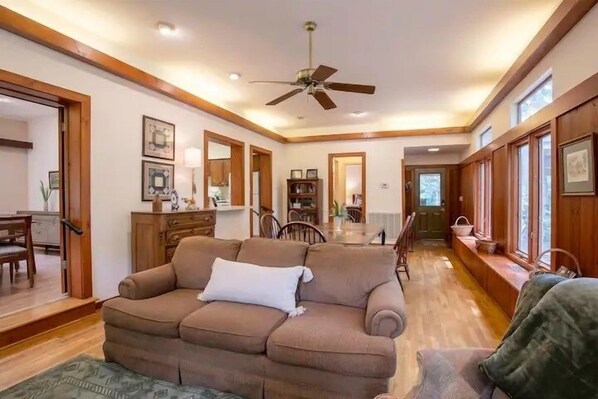 A cozy living room with a brown sofa, wooden walls, a ceiling fan, and a dining area in the background.