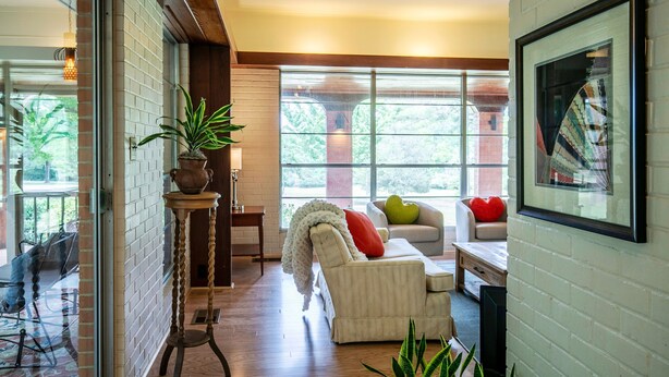 Una sala de estar moderna con pared de ladrillo blanco, un sofá con cojines rojos y verdes, una mesa de centro y un ventanal con vistas a follaje exterior.