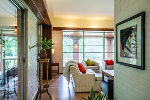 A modern living room with a white brick wall, a sofa with red and green cushions, a coffee table, and a large window offering a view of greenery outside. - The Shields, Family Stay by Velvet Ditch Villas (Oxford)