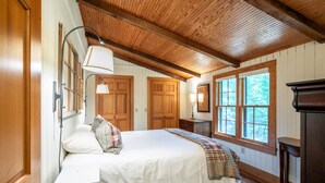 A bedroom with a wooden ceiling, a bed with a white bedspread, a bedside lamp, and a window with a view of trees. - The Lil' Sipp, 2BR Cabin by Velvet Ditch Villas (Oxford)