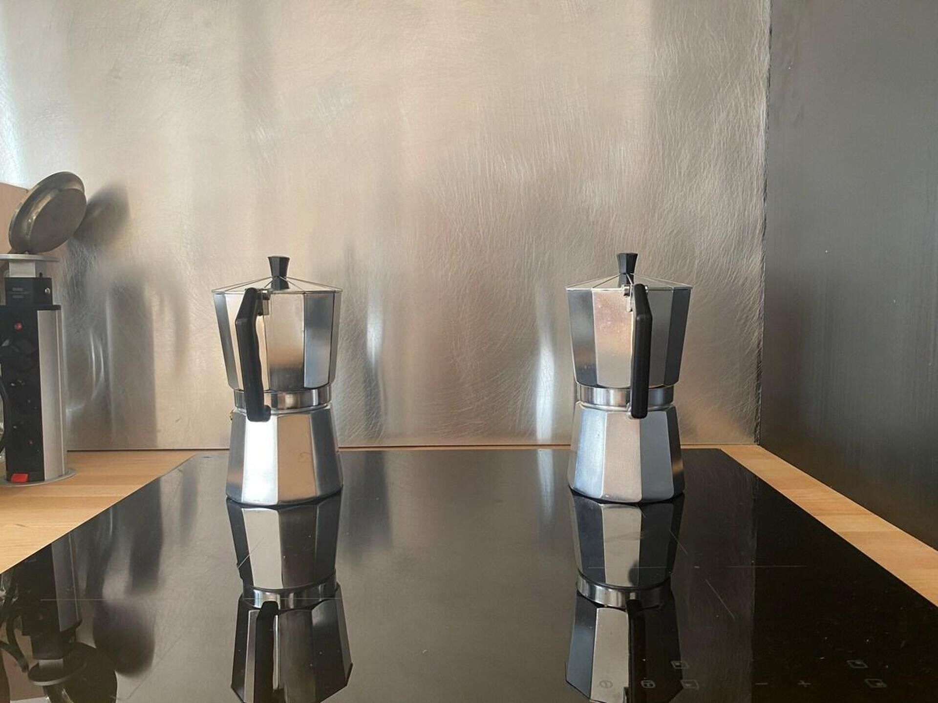 Zwei Edelstahl-Kaffeemaschinen auf einer spiegelnden schwarzen Oberfläche vor einer schlichten Wand.
