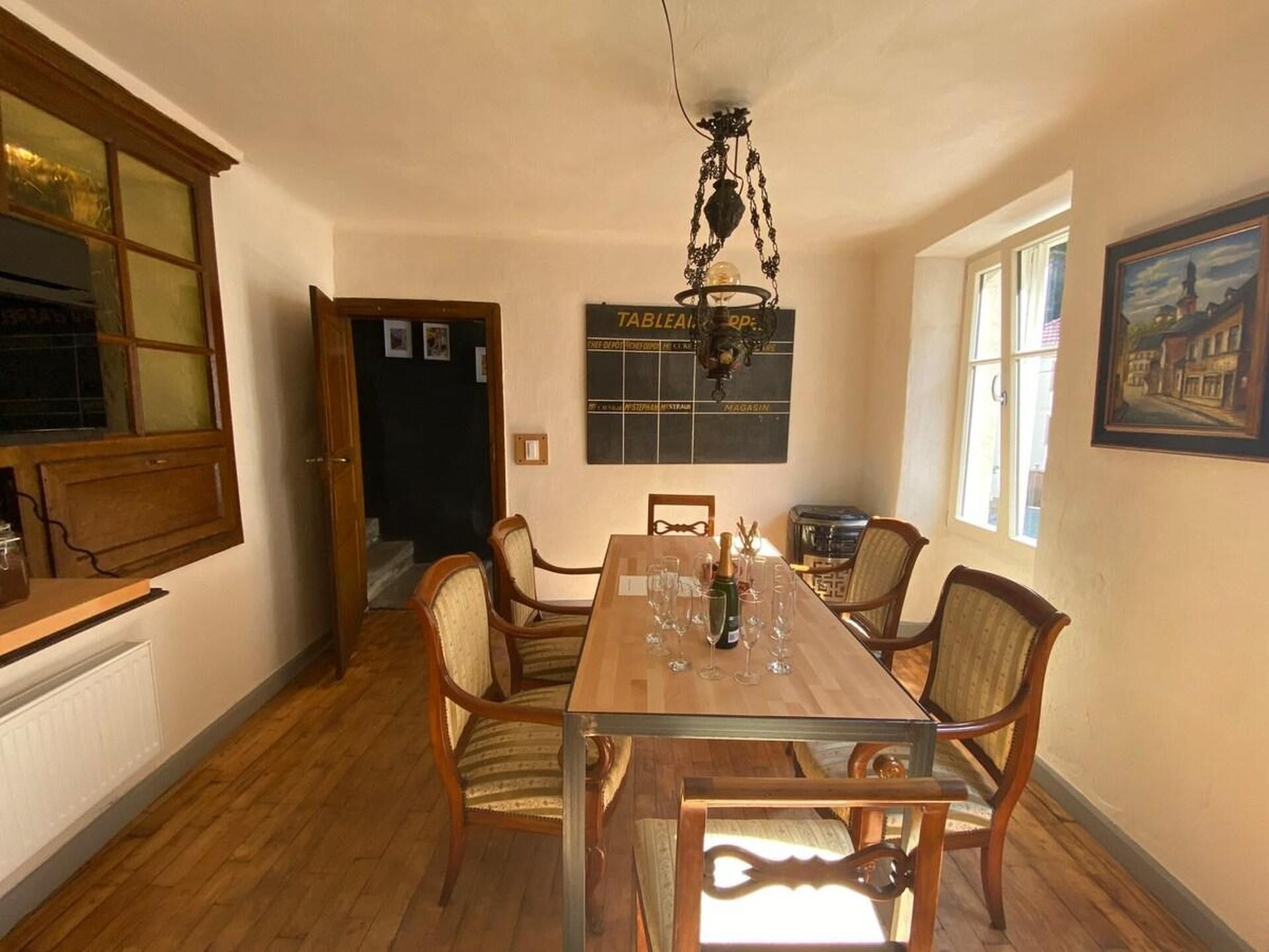 Un coin repas avec une table en bois, des chaises et un lustre. Il y a une fenêtre donnant sur un immeuble et un tableau accroché au mur.