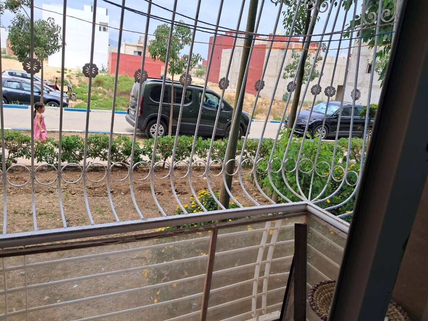 Vista a través de una reja metálica que da a un área pavimentada, un niño caminando y coches estacionados.