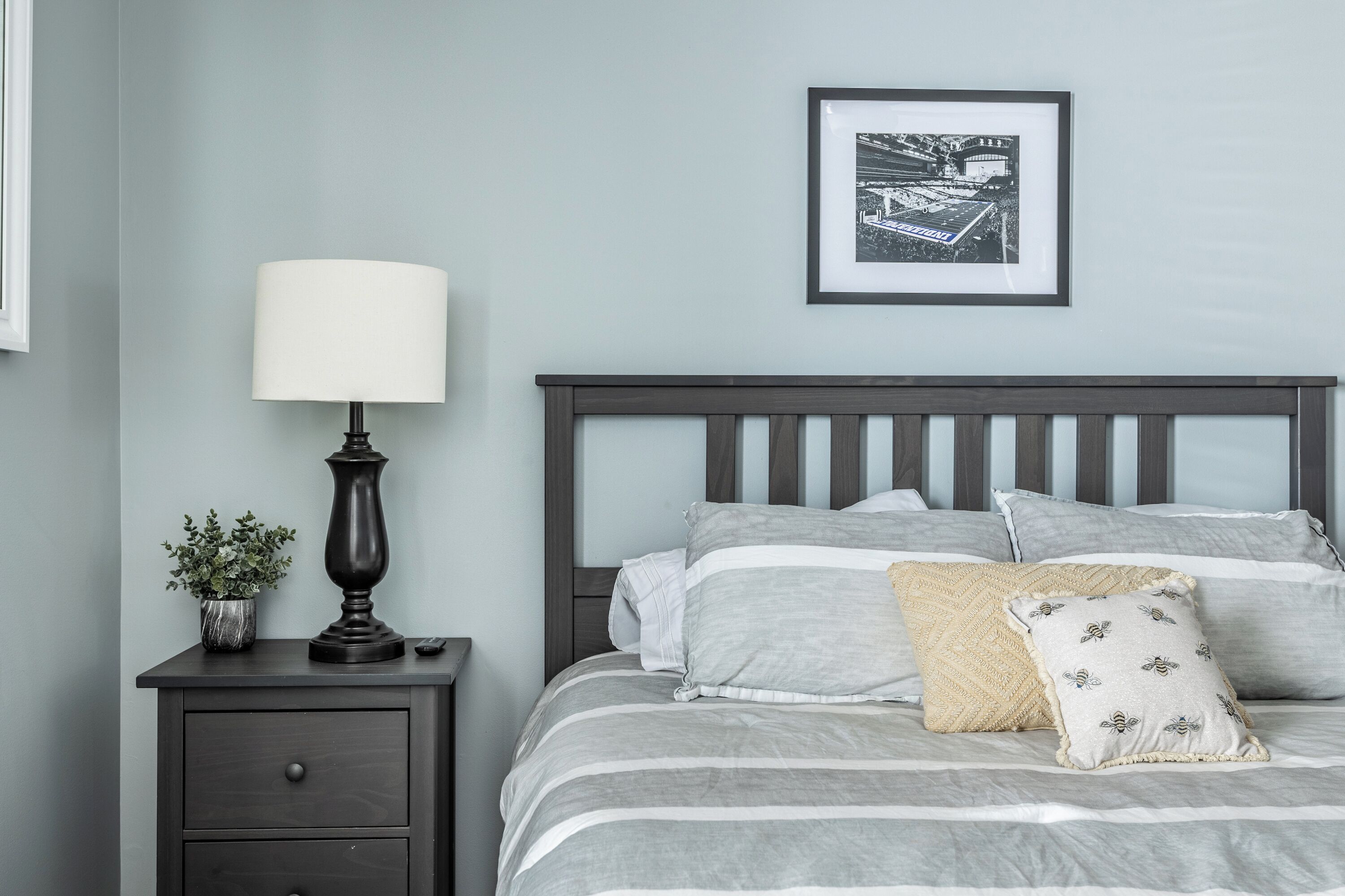 A neatly made bed with striped bedding, a bedside lamp, a framed picture on the wall, and a small plant on a nightstand.