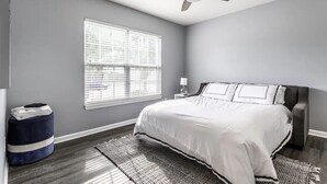 Quarto com cama, ventilador de teto, janela com persianas, tapete e mesinha de cabeceira com abajur.