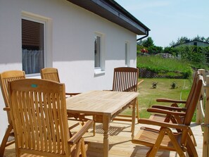 A wooden deck with a table and chairs, a white building with windows, and a garden in the background. - Ferienhaus in Bansin am See (Heringsdorf)