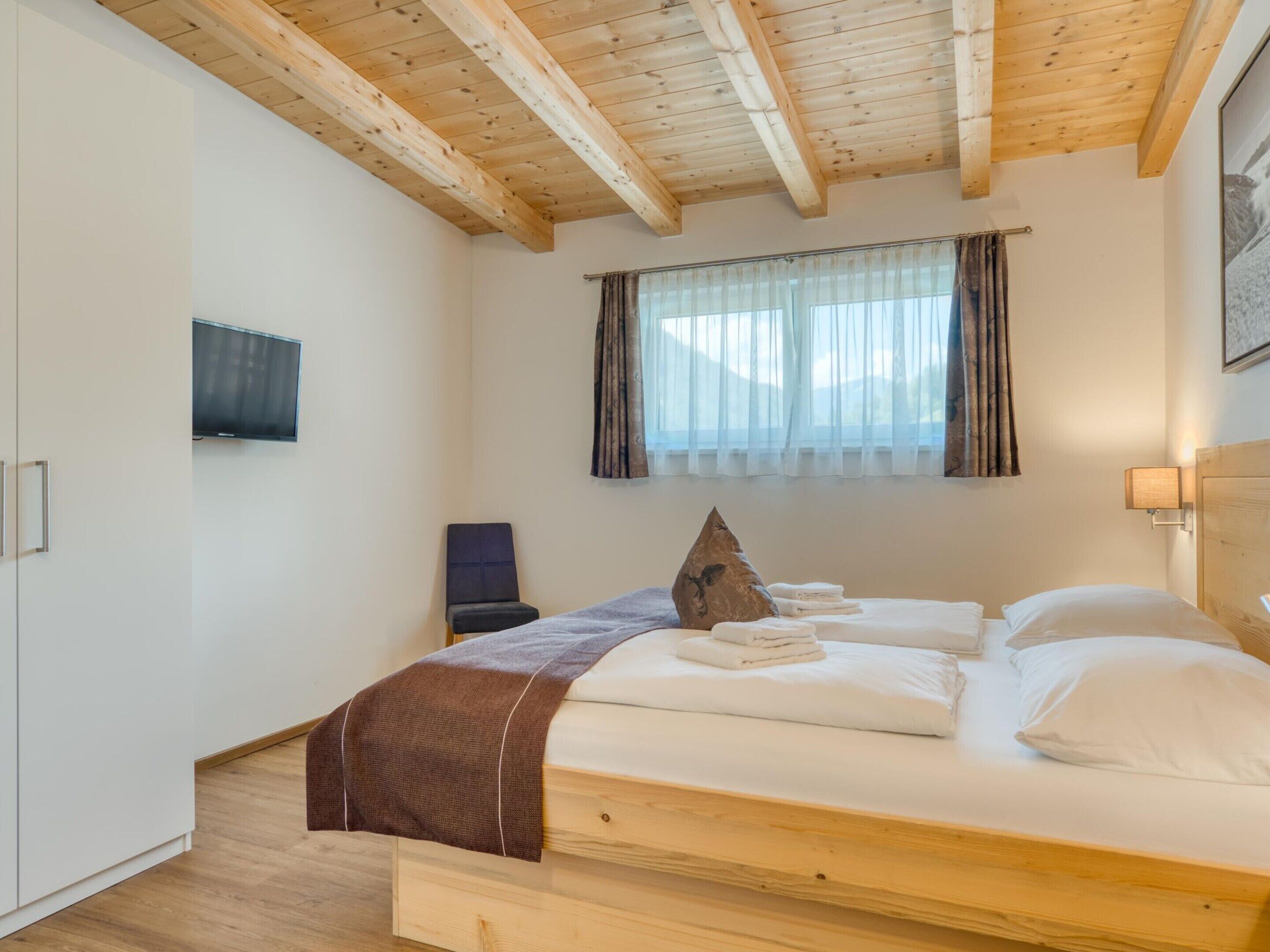 Een slaapkamer met een houten plafond, een bed met witte lakens, een aan de muur bevestigde televisie en een raam met gordijnen.
