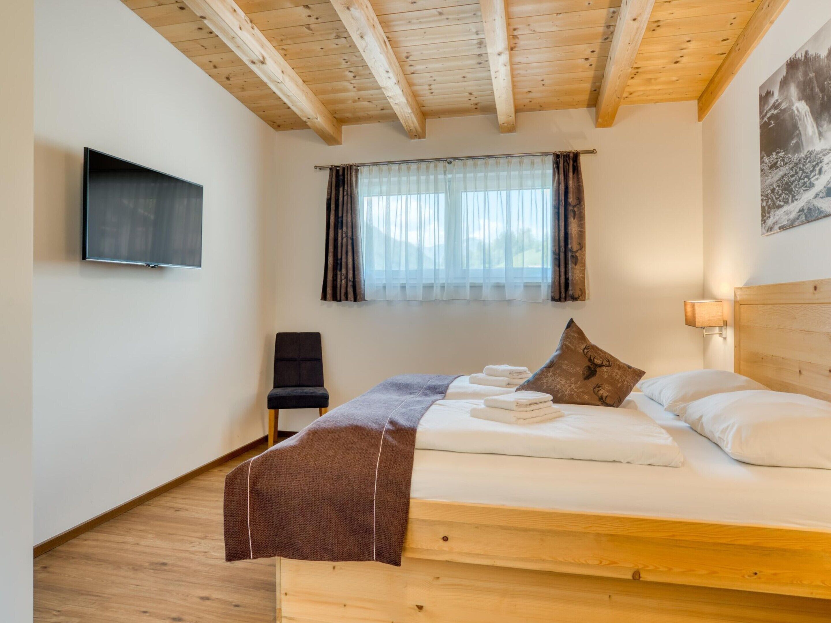 A bedroom with a wooden ceiling, a bed with white linens, a chair, a TV mounted on the wall, and a window with curtains.
