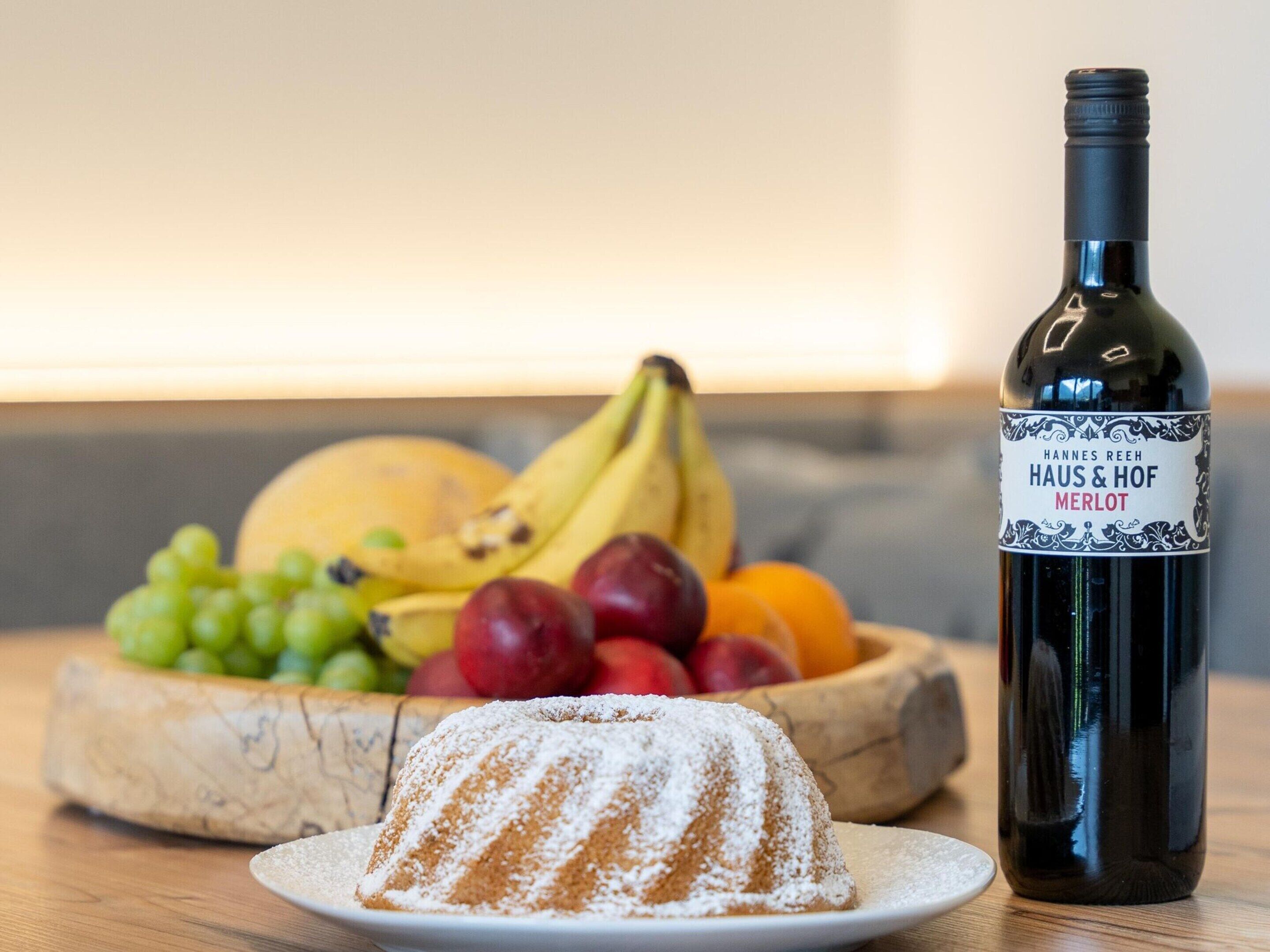 A bottle of Hannes Reeh Haus & Hof Merlot wine, a fruit bowl with bananas, grapes, and apples, and a dusted bundt cake on a plate.