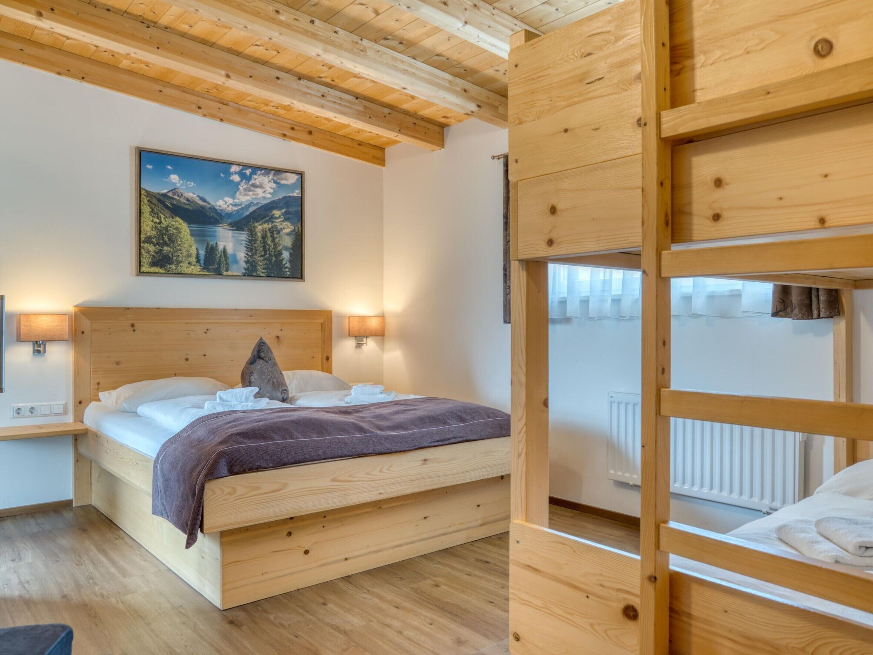 A wooden bunk bed room with a painting of a mountain landscape, a bedside table with a lamp, and a radiator.