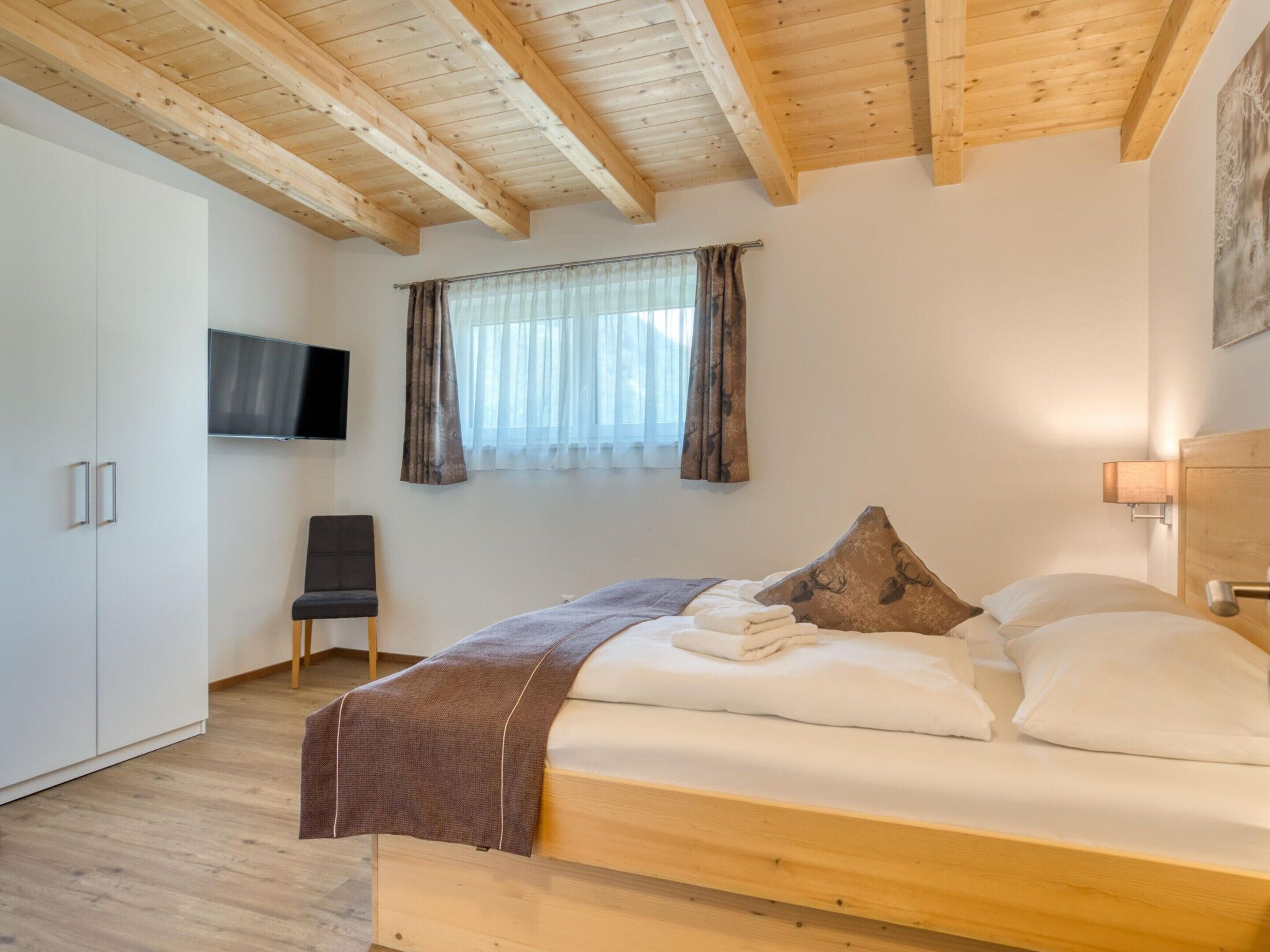Un dormitorio con una cama de madera, televisor montado en la pared, una silla y un armario.