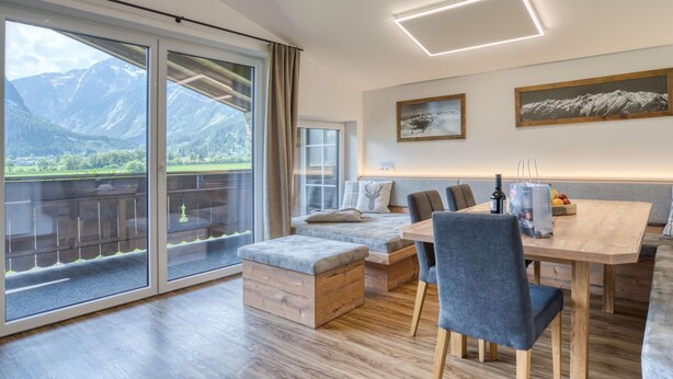 A modern dining area with a wooden table, grey upholstered chairs, and a bench. A large window offers a view of mountains and a green landscape. - Holiday Home in Neukirchen With Mountain Views (Neukirchen am Grossvenediger)