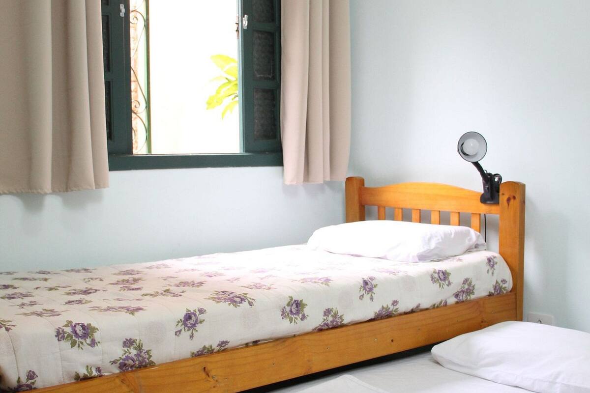 Ein Einzelbett mit Holz-Kopfstück, ein Nachttisch mit Lampe und ein Fenster mit Vorhängen.