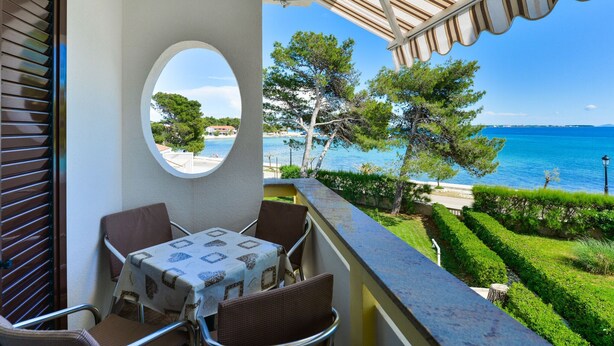 Un balcón con mesa y sillas, con vista al mar azul y un jardín cuidado.