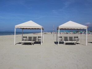Två tält placerade vid stranden, med solstolar, en klar himmel och en avlägsen utsikt över stranden och havet.