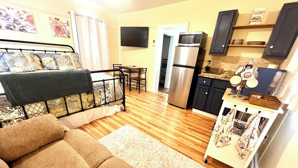 A compact living space with a sofa, a small dining table, a kitchen area with a refrigerator and microwave, and a TV mounted on the wall. - Historic Hilltop Guesthouse - Charming studio/cottage one block from downtown  (Laurel)