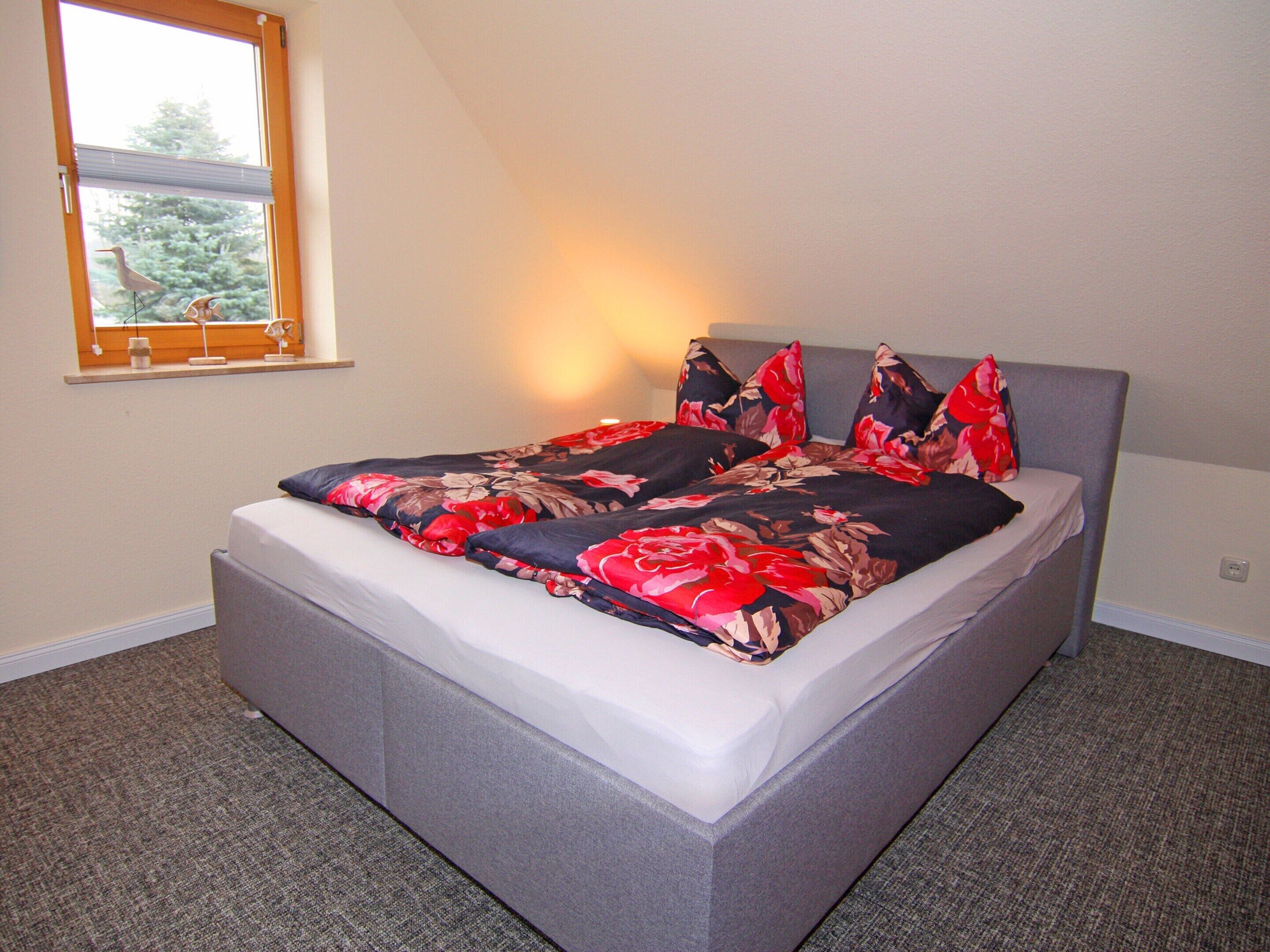 A bed with a floral bedspread and multiple pillows, a window with blinds, and a grey upholstered headboard.