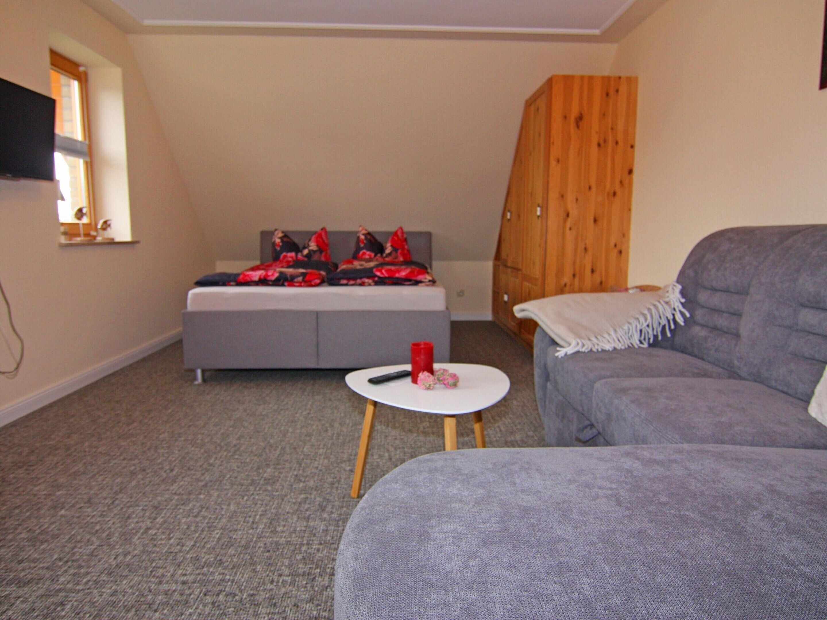 A room with a grey sofa, a small round table with a red cup, a grey bed with red and black pillows, and a wooden wardrobe.