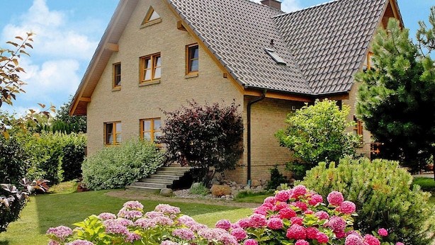 Una casa de dos pisos con techo inclinado, fachada de ladrillo y un jardín delantero con flores rosadas y arbustos verdes.