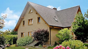 A two-story house with a sloped roof, brick exterior, and a front garden with pink flowers and green shrubs.