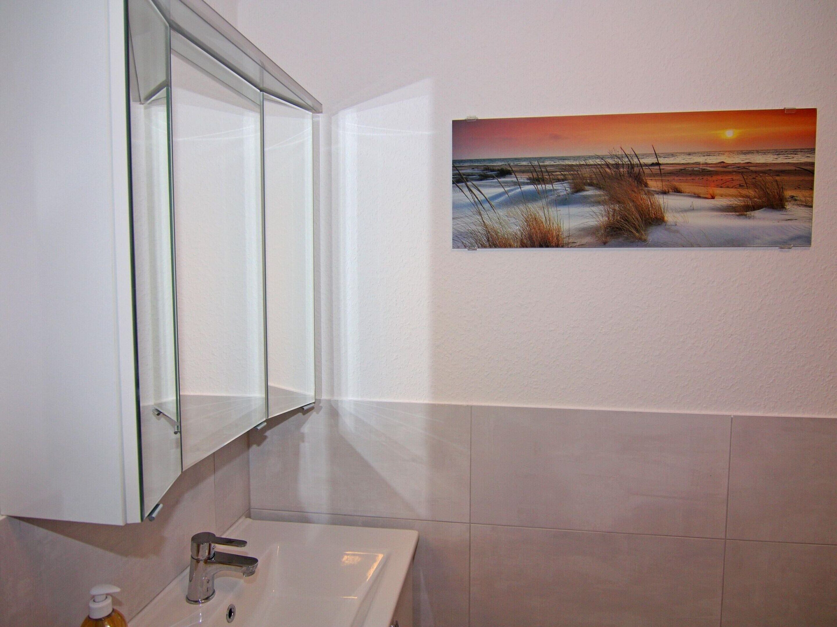 A bathroom with a white sink, a glass shower enclosure, and a wall-mounted painting of a sunset over a beach.