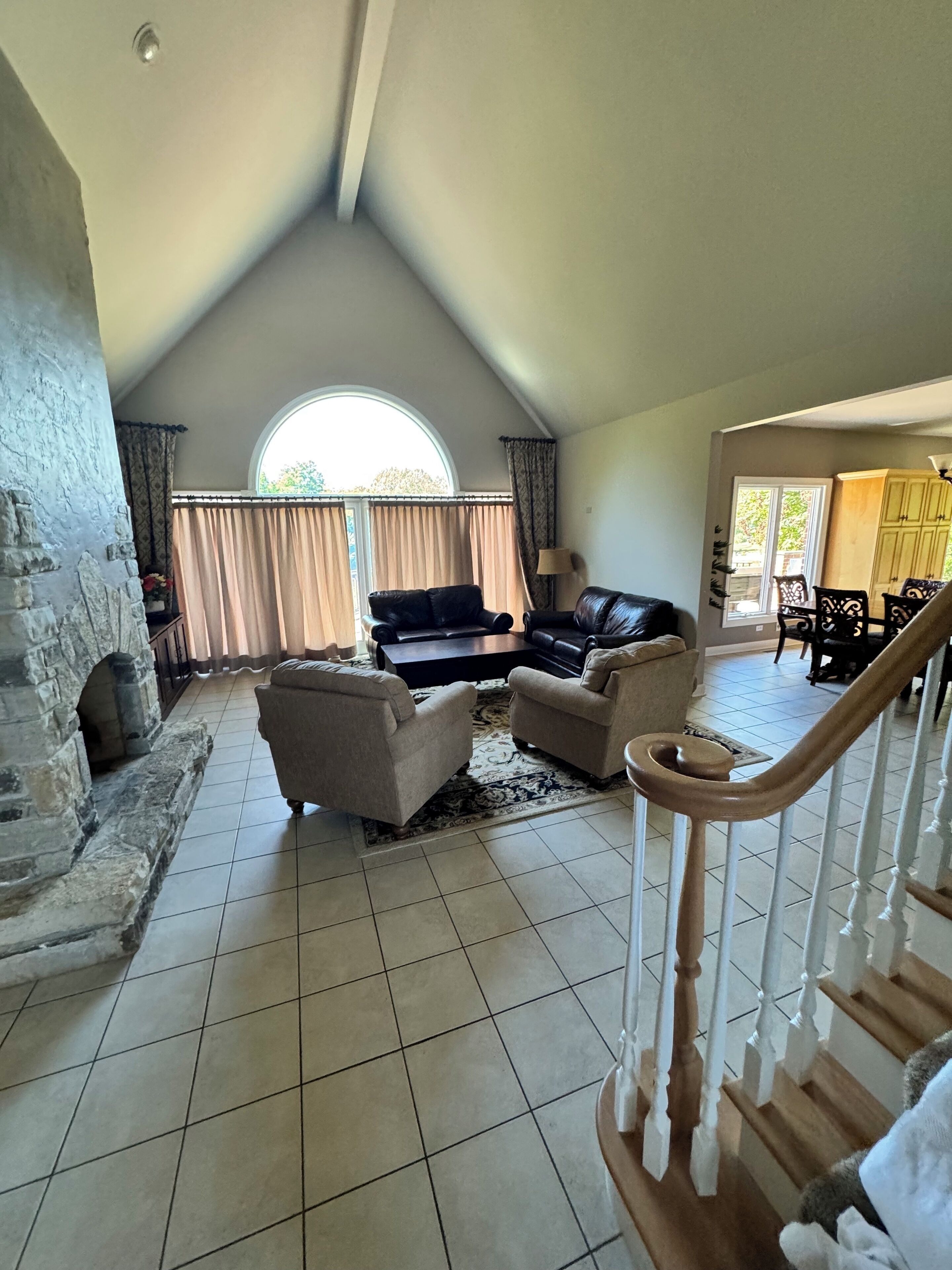 A spacious living room with a stone fireplace, a black leather sofa, beige armchairs, and a wooden staircase.