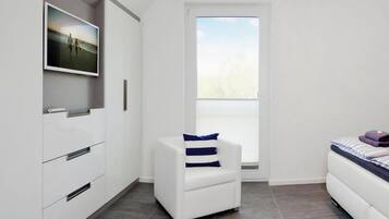 Un dormitorio moderno con una cama, un sillón blanco, un televisor montado en la pared y un ventanal grande.
