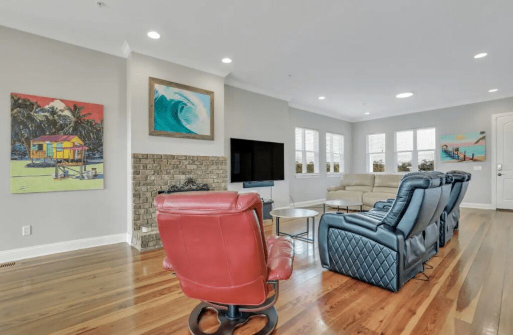 A spacious living room with hardwood floors, a red leather chair, a blue massage chair, a flat-screen TV, and artwork on the walls.
