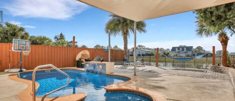 An outdoor swimming pool with a basketball hoop, palm trees, and a view of a house in the background.