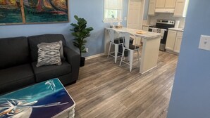 A living room with a sofa, a coffee table, and a painting on the wall. There is a kitchen in the background with white cabinets and a dining area with a table and chairs. - Captain & Mermaid's Diamond #BP25 (Big Pine Key)