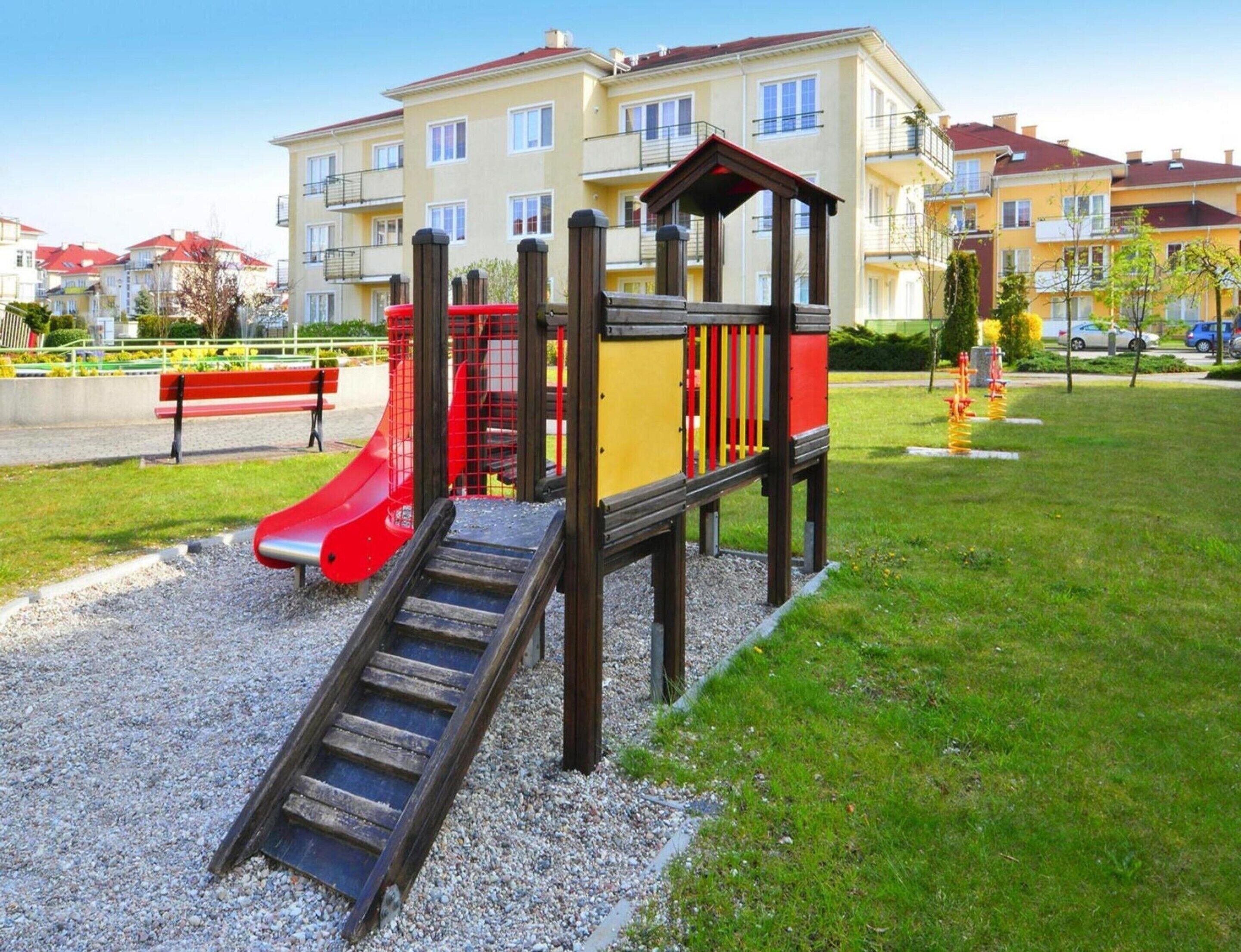 A playground with a slide and climbing structure, surrounded by a grassy area and residential buildings in the background.