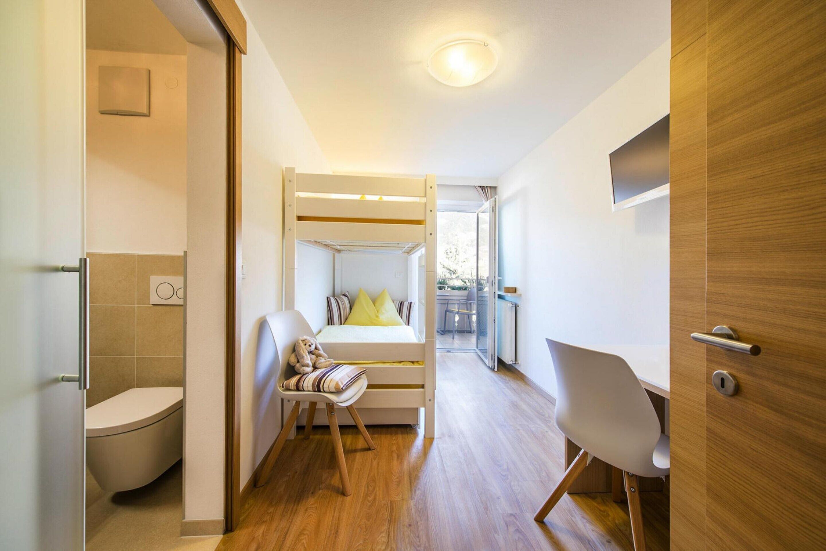 A compact, modern room with a wooden floor, a bunk bed with a desk and chair, a white toilet, and a small window.