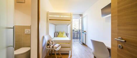 A compact, modern room with a wooden floor, a bunk bed with a desk and chair, a white toilet, and a small window.