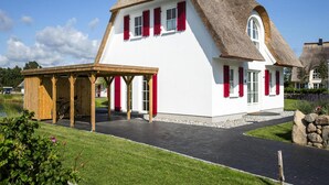 A white house with a thatched roof and red shutters, a wooden pergola, and a paved driveway. - Luxushaus Boddenkuste mit Sauna (Fuhlendorf)