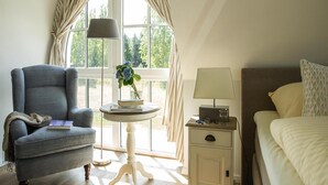 A hotel room with a blue armchair, a bedside table with a lamp, a small table with a vase of flowers, and a bed with a patterned pillow. - Luxushaus Boddenkuste mit Sauna (Fuhlendorf)