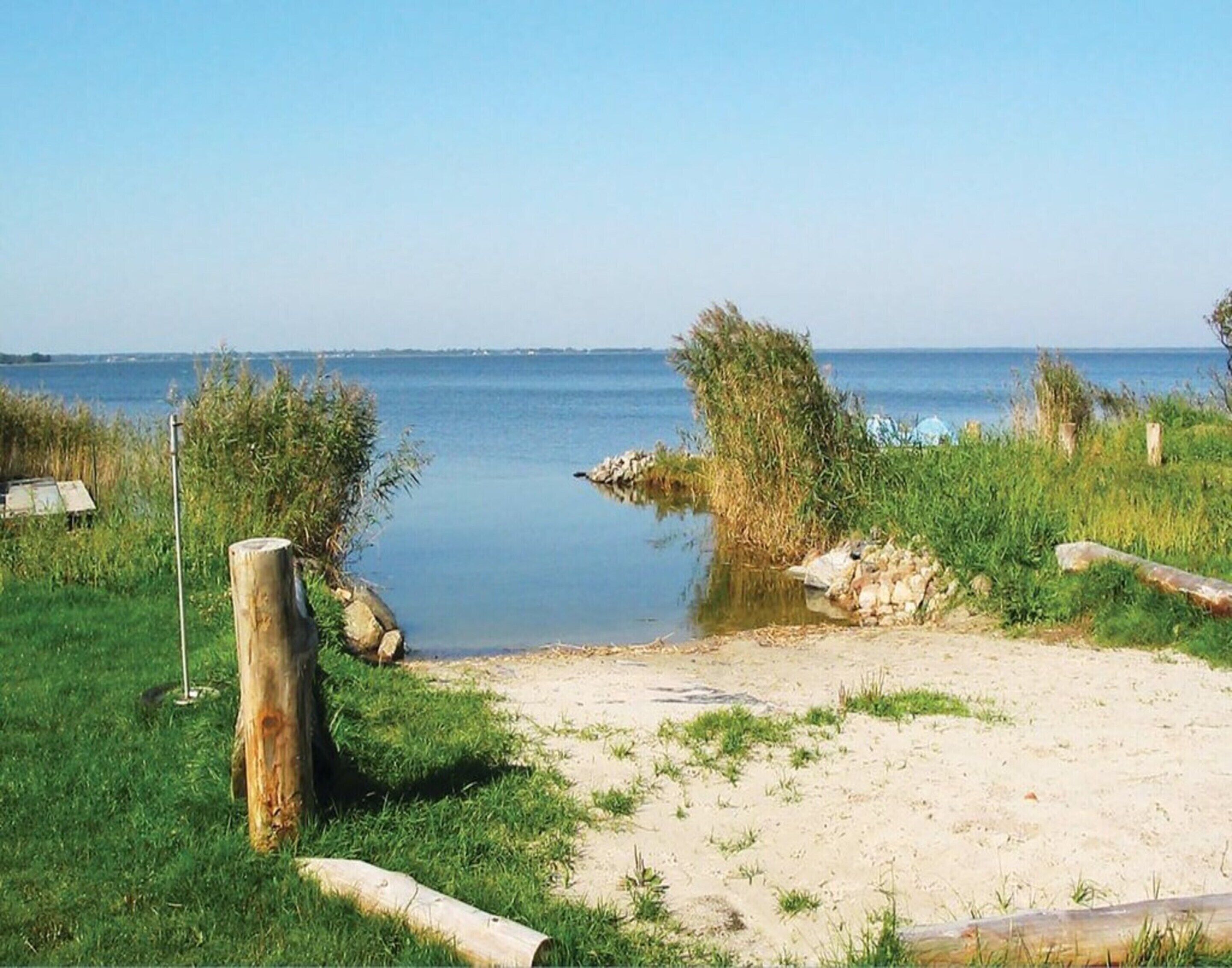 A sandy beach with a calm body of water, surrounded by grassy areas and a wooden fence.