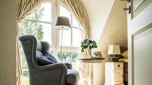 A cozy room with a grey armchair, a small table with a lamp, and a view of greenery through large windows. - Luxushaus Boddenkuste mit Sauna (Fuhlendorf)