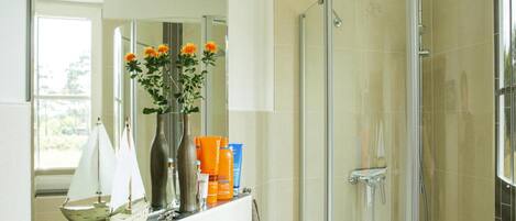 A modern bathroom with a glass shower enclosure, a vanity with toiletries, and a large mirror reflecting a sailboat model and flowers.