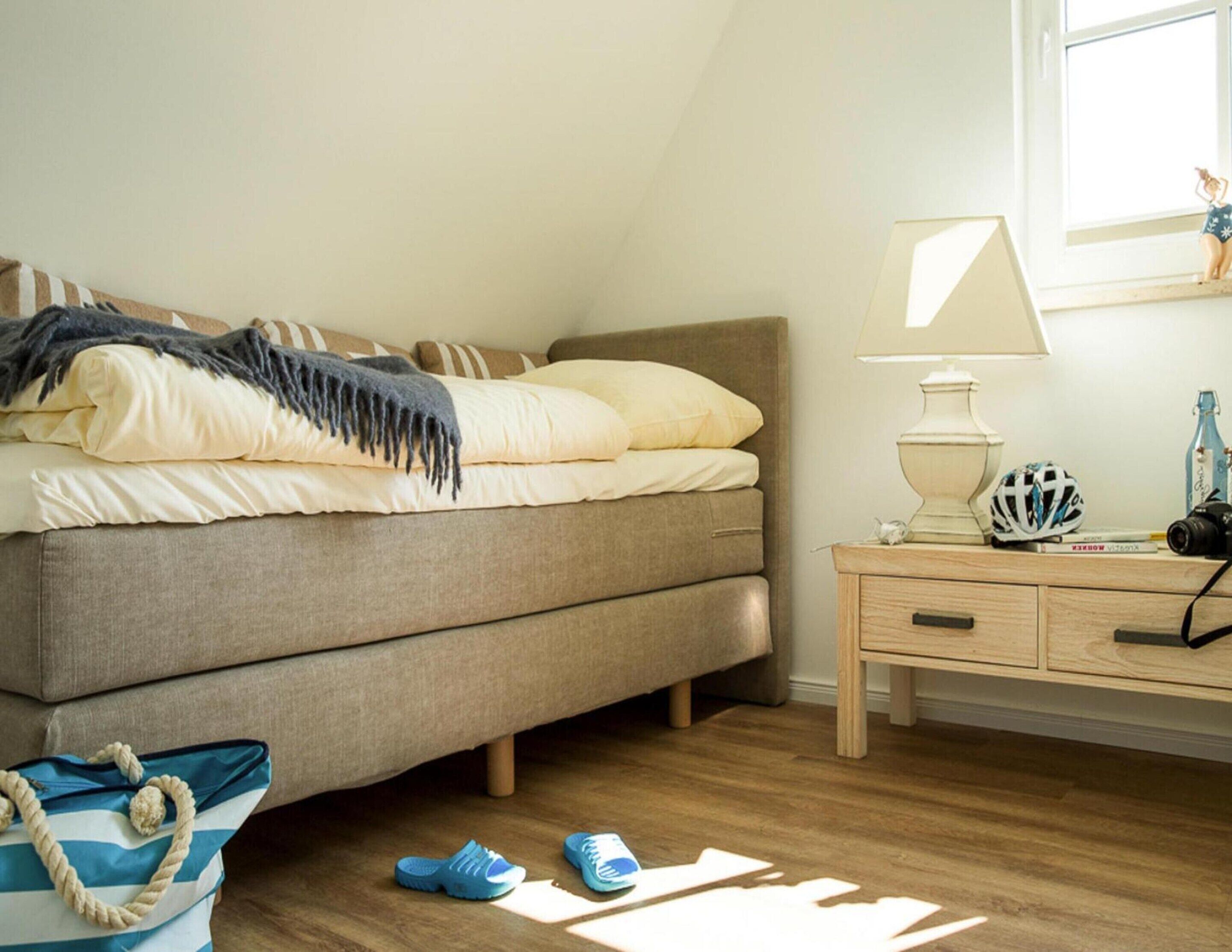 Une chambre à coucher avec un lit, une table de chevet, une lampe et un sac contenant des chaussons posé sur le sol.