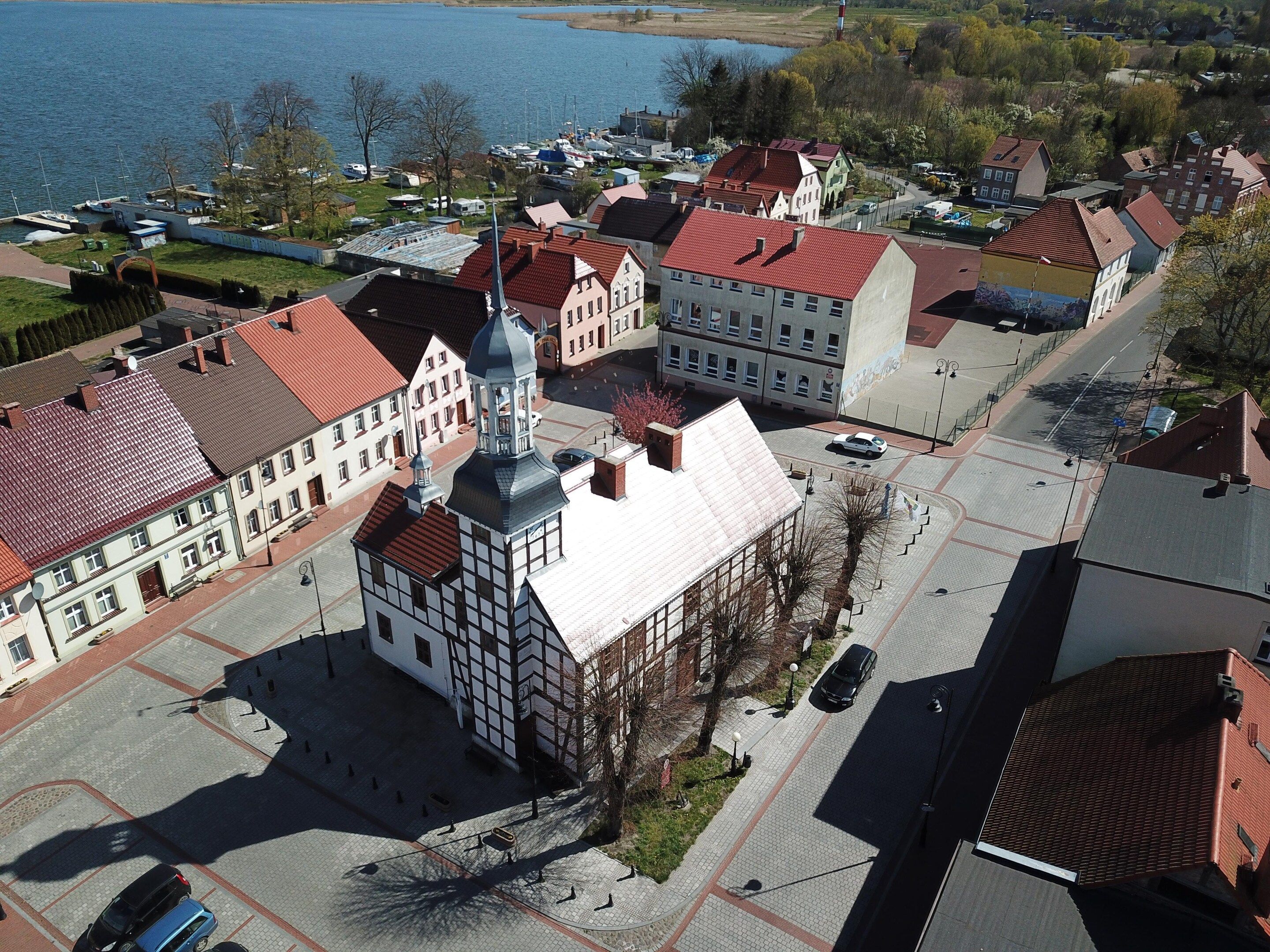 Une place de ville bordée de bâtiments historiques, avec une tour centrale et un accès au bord de l’eau.