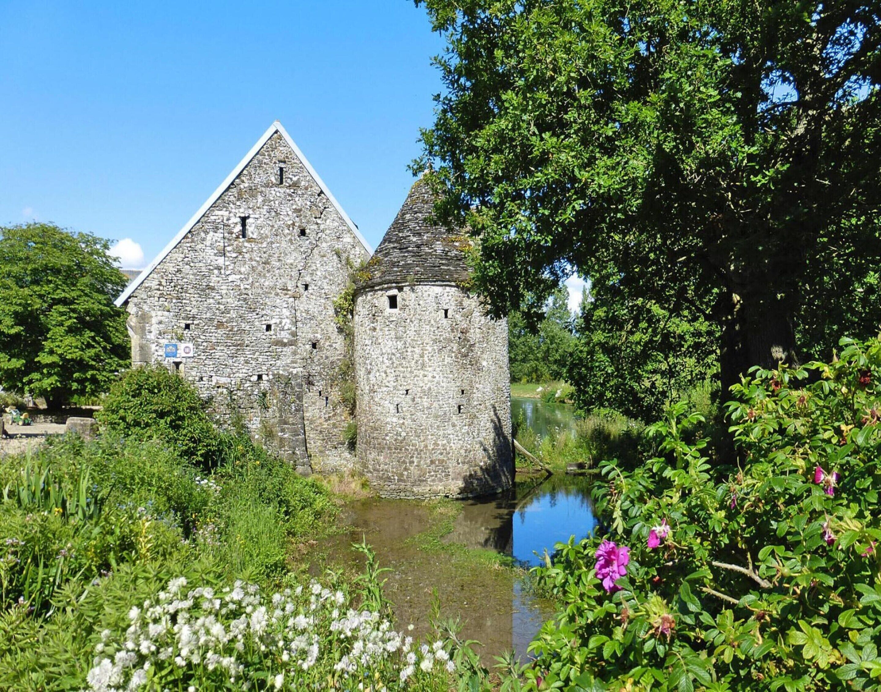 Un bâtiment en pierre doté d’un toit triangulaire et d’une tour cylindrique, entouré d’une végétation luxuriante et bordé d’une étendue d’eau paisible.