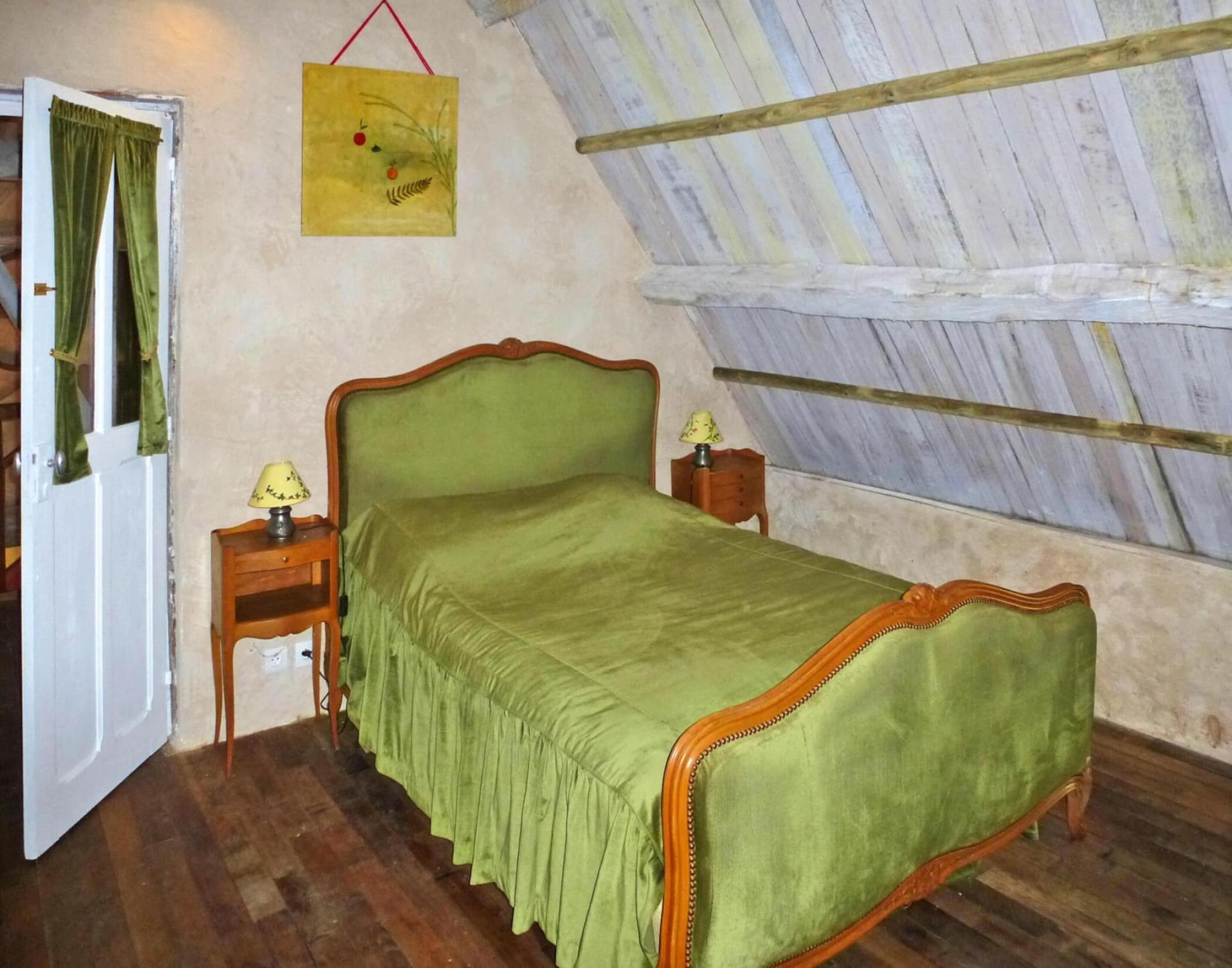 Une chambre avec un lit recouvert d’un tissu vert, une table de chevet en bois avec une lampe, et une petite table également dotée d’une lampe. Un tableau est accroché au mur, et la pièce a un plafond en pente.