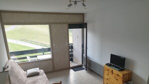 A room with a sofa, a table, a TV on a wooden stand, a dresser, and a sliding glass door leading to an outdoor area. - Studio apartment at the foot of the slopes (St Aventin)