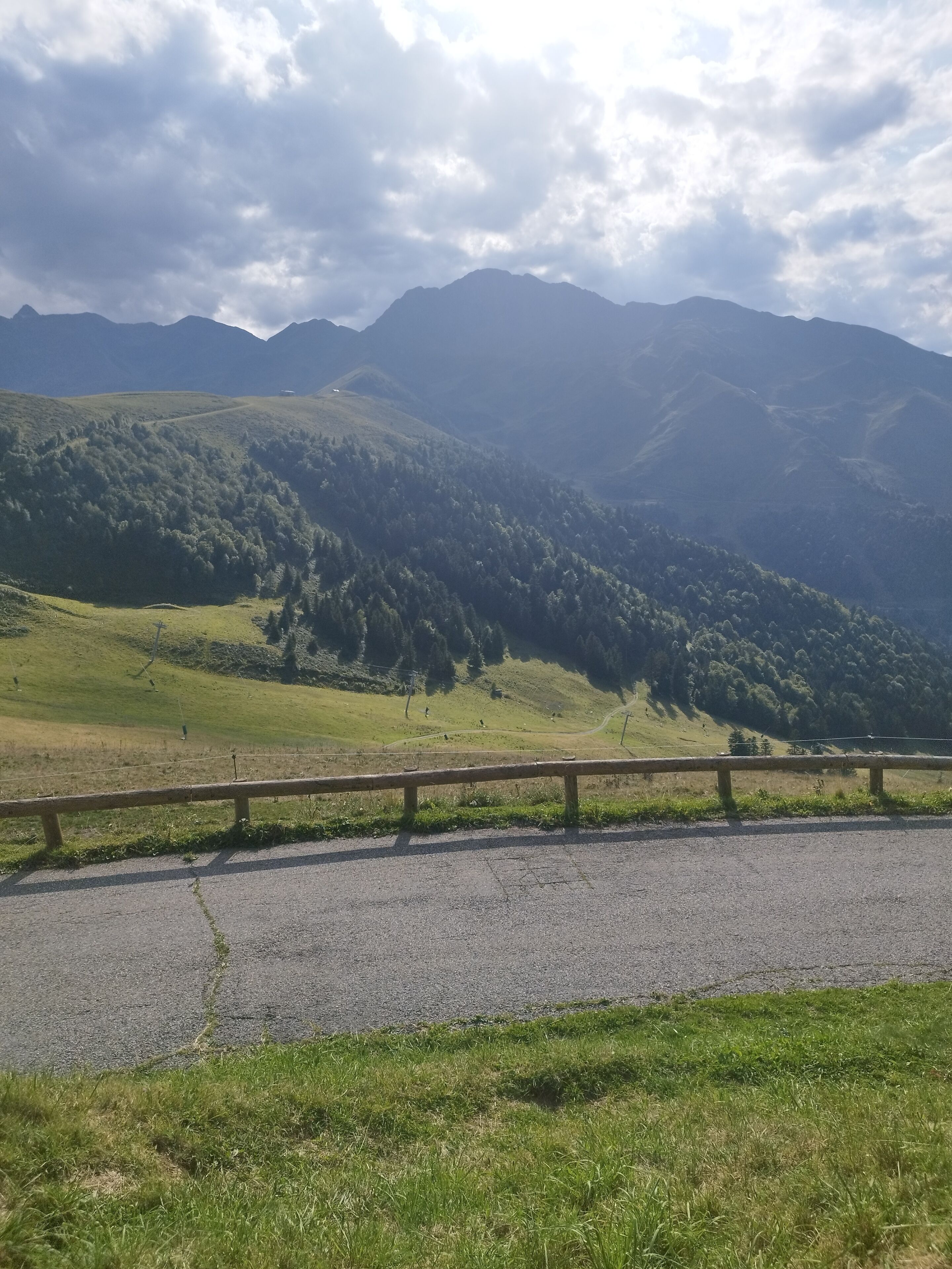 A paved road with a guardrail, surrounded by lush green hills and mountains under a partly cloudy sky.