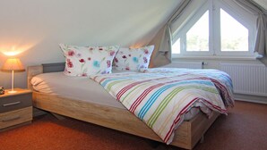 A bedroom with a wooden bed, floral pillows, a striped blanket, a nightstand with a lamp, and a window with curtains. - Appartementhaus Strandloper, Zinnowitz (Zinnowitz)