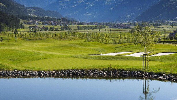 A golf course with a pond, mountains in the background, and a small village. - Apartment in Fugen Near Spieljochbahn (Fuegen)