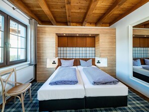 Une chambre double avec deux lits, des poutres apparentes en bois au plafond, une tête de lit à carreaux, des lampes de chevet, une chaise et une fenêtre avec des rideaux.